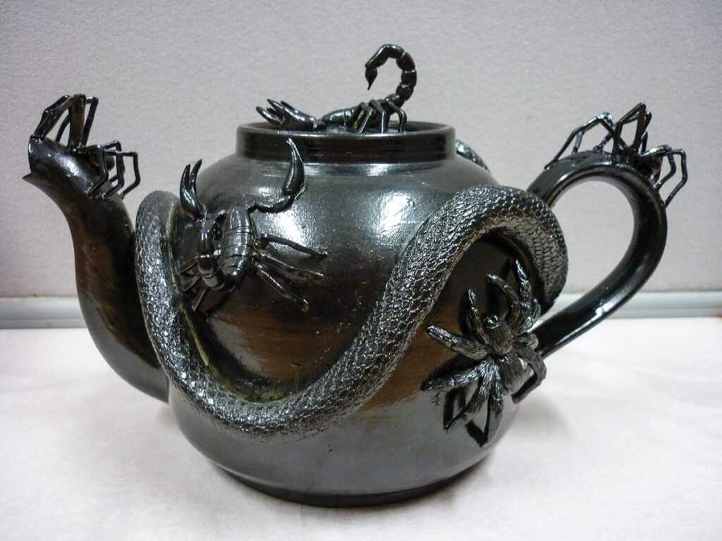 The Black Teapot made by Roger Law for the “Teapotmania” exhibition in Norwich in 1995, which told the story of the British craft teapot and teacosy. The teapot and lid are decorated with an applied moulded snake, three spiders, and two scorpions, then spray-painted black.